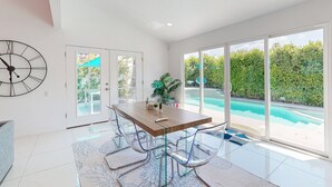 Un comedor moderno con una mesa de madera y sillas de vidrio transparente, ventanas amplias con vistas a una piscina, y un reloj en la pared.