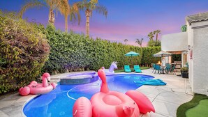 Une piscine avec des jeux gonflables, un espace patio avec des chaises et une table, et une vue sur les palmiers.