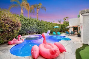 A swimming pool with inflatable toys, a patio area with chairs and a table, and a view of palm trees. - Desert Delight | Corner Lot, Pool & Spa | Putting Greens & Hammock (Palm Desert)
