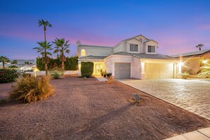 A two-story house with a garage, surrounded by landscaped gardens and palm trees. - Desert Delight | Corner Lot, Pool & Spa | Putting Greens & Hammock (Palm Desert)