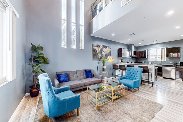Ein geräumiges Wohnzimmer mit grauem Sofa, blauen Sessel und Couchtisch. Ein großes Fenster lässt viel Tageslicht herein, und eine Pflanze sorgt für Grün.