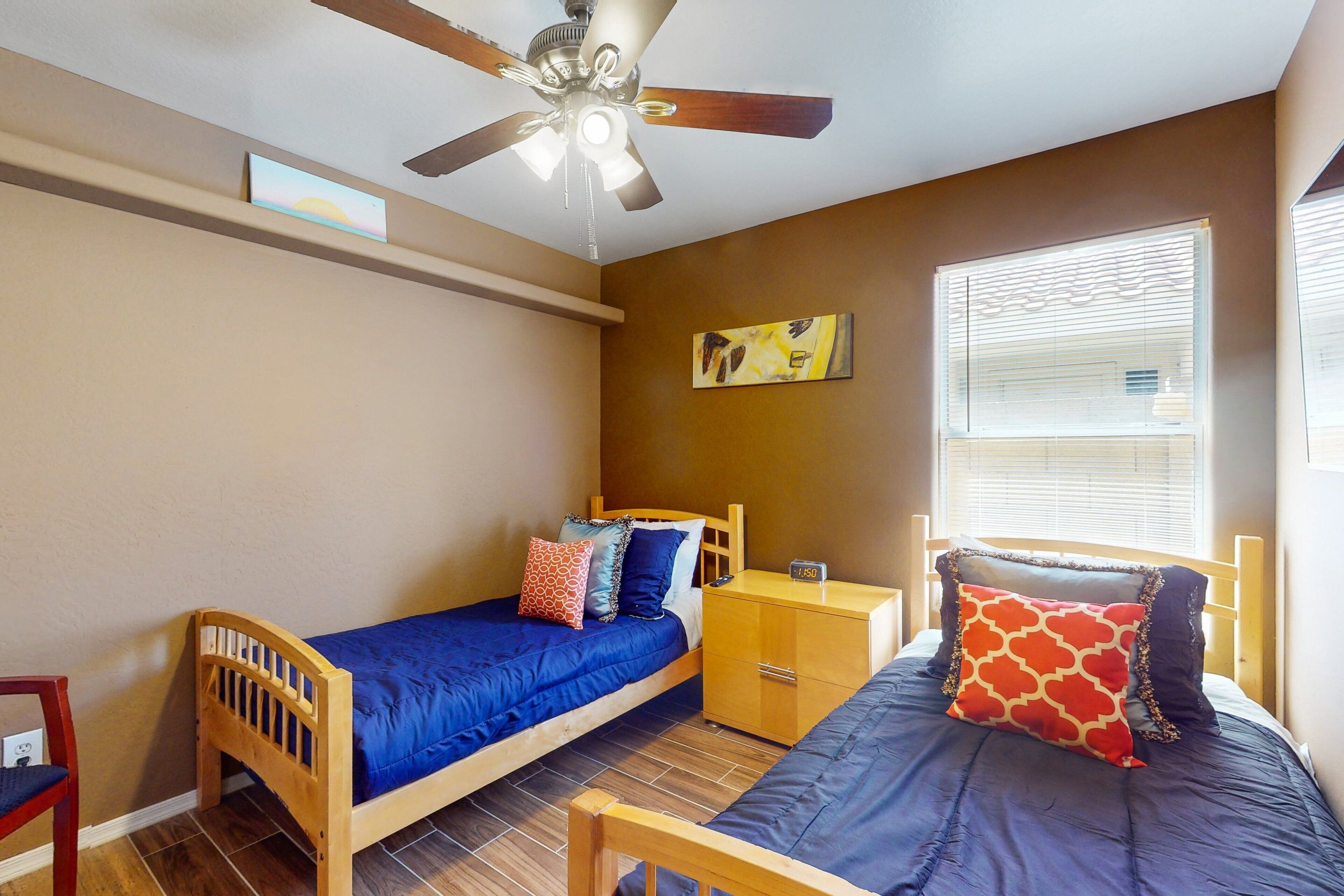Une chambre avec deux lits simples, une table de chevet en bois, un ventilateur de plafond et une fenêtre avec des stores.