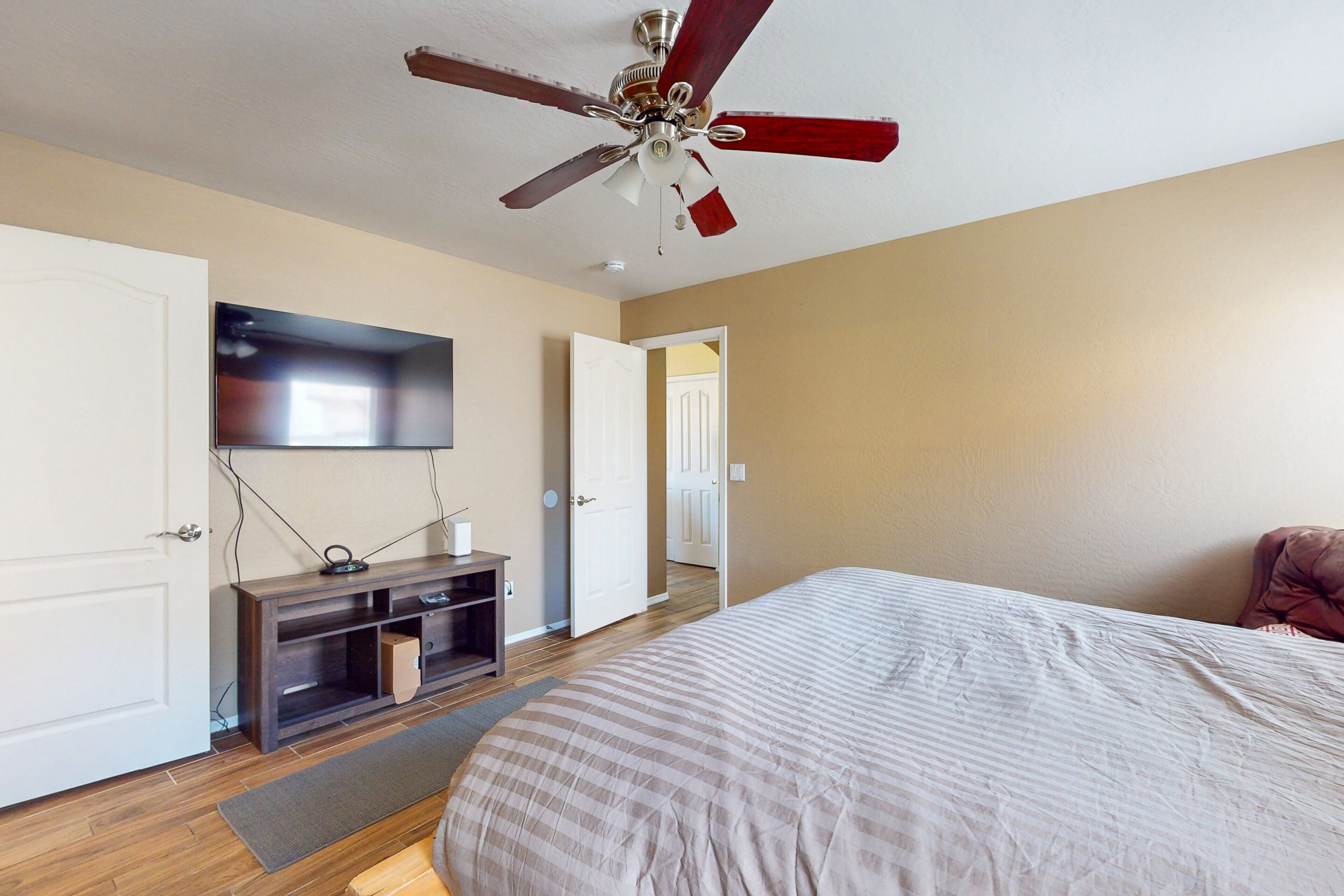 A bedroom with a bed, a ceiling fan, a TV mounted on the wall, and a door leading to another room.
