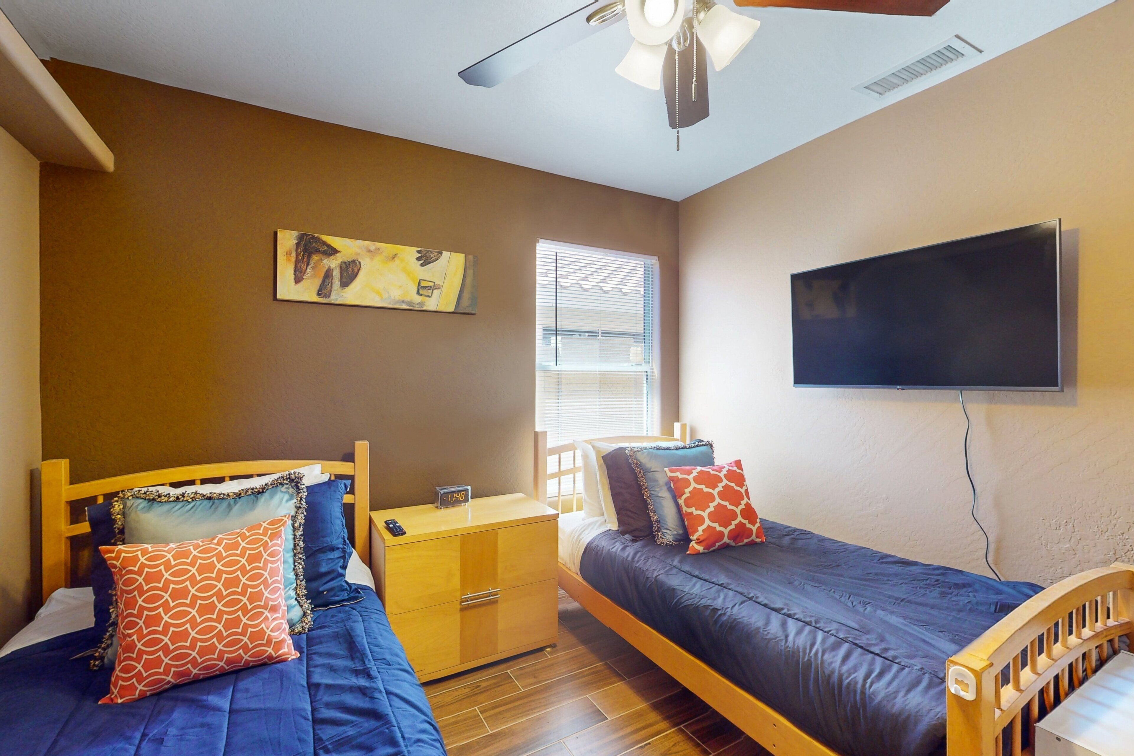 A bedroom with two beds, a nightstand, a wall-mounted TV, and a ceiling fan.
