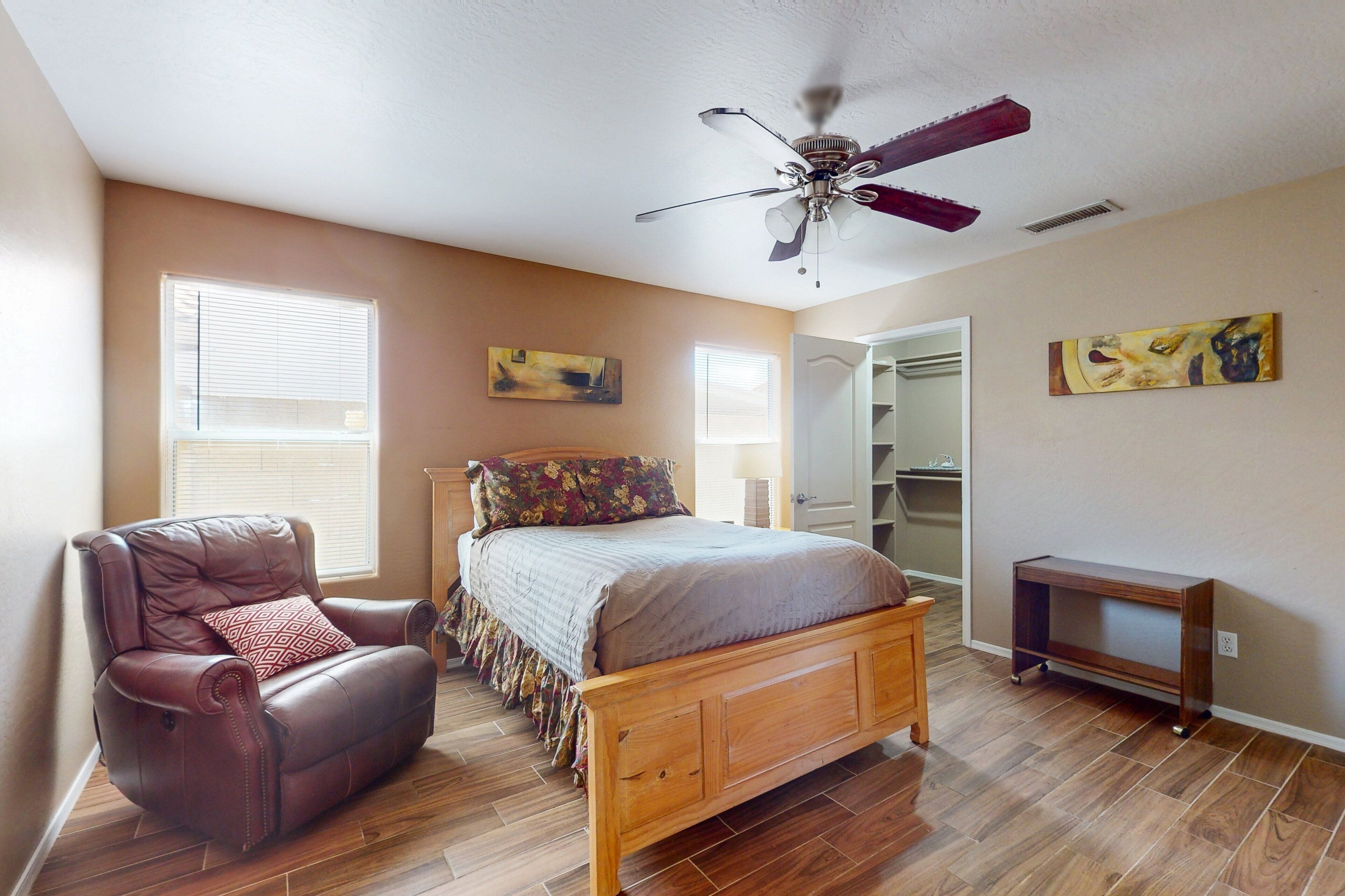 A bedroom with a wooden bed, a leather chair, a ceiling fan, and two windows.