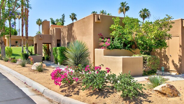 A desert-style building with stucco walls, a landscaped garden with desert plants and pink flowers, and palm trees in the background. - Cozy Dog-Friendly Condo, Shared Pool/Hot Tub, Near Golf Courses & National Park (Palm Desert)