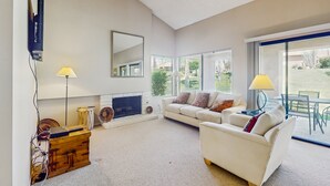 A spacious living room with a fireplace, a sofa set, a television mounted on the wall, and a view of the outdoors through large windows. - Cozy Dog-Friendly Condo, Shared Pool/Hot Tub, Near Golf Courses & National Park (Palm Desert)