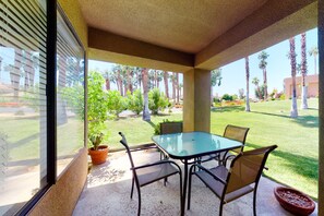 Eine überdachte Terrasse mit Glastisch und Stühlen, einem Blumentopf und Blick auf einen gepflegten Rasen und Palmen.