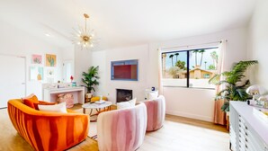 A modern living room with a fireplace, a large window with a view of palm trees, and a bright chandelier. - Dog-friendly desert dream with a private pool, pool spa, & putting green (Palm Springs)