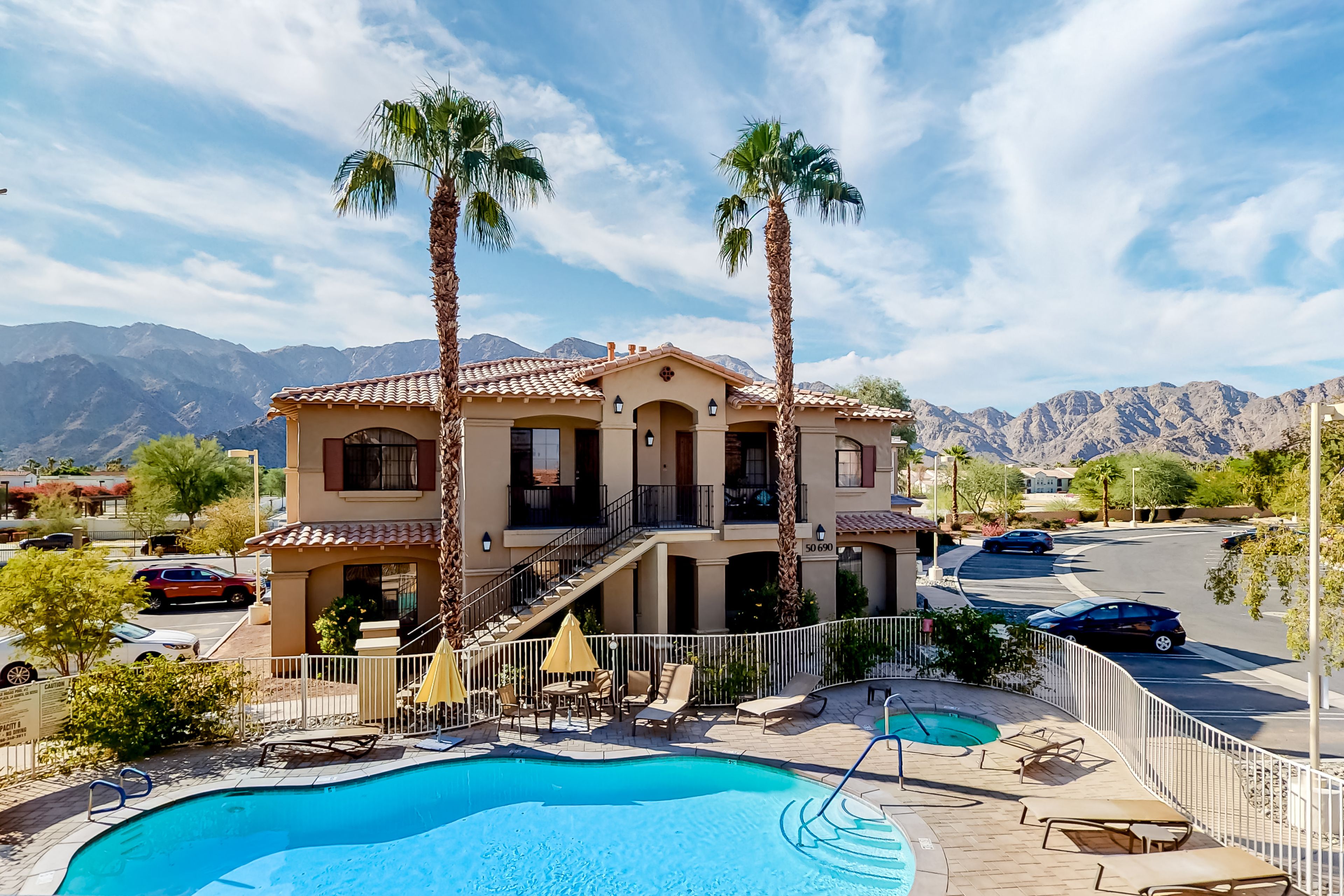 A two-story building with a swimming pool, palm trees, and mountains in the background.
