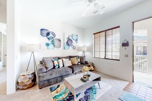 A living room with a grey sofa, two wall-mounted lamps, abstract art, a coffee table, and a view through a window with blinds. - 2nd-Floor Condo with Central AC, Shared Pool - ​#LIC-260042 (La Quinta)