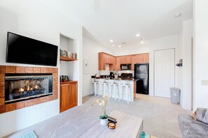 A modern living room with a fireplace, a flat-screen TV, and a kitchen area with white bar stools. - 2nd-Floor Condo with Central AC, Shared Pool - #LIC-260042 (La Quinta)