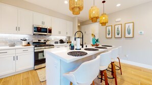 Une cuisine moderne avec des meubles blancs, un îlot central doté de tabourets de bar, un four à micro-ondes, une cuisinière et des lustres suspendus en osier.