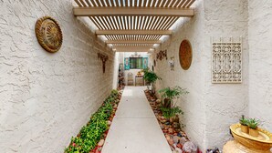 Ein schmaler Korridor mit Fliesenboden, weißen Wänden und einer Holzpergola darüber. An den Wänden befinden sich dekorative Teller und weiter hinten ein kleiner Tisch mit Stühlen.