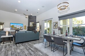 Un salón moderno con un sofá gris, una mesa de comedor y sillas. Hay una chimenea, obras de arte en las paredes y un gran ventanal con vistas al verdor.