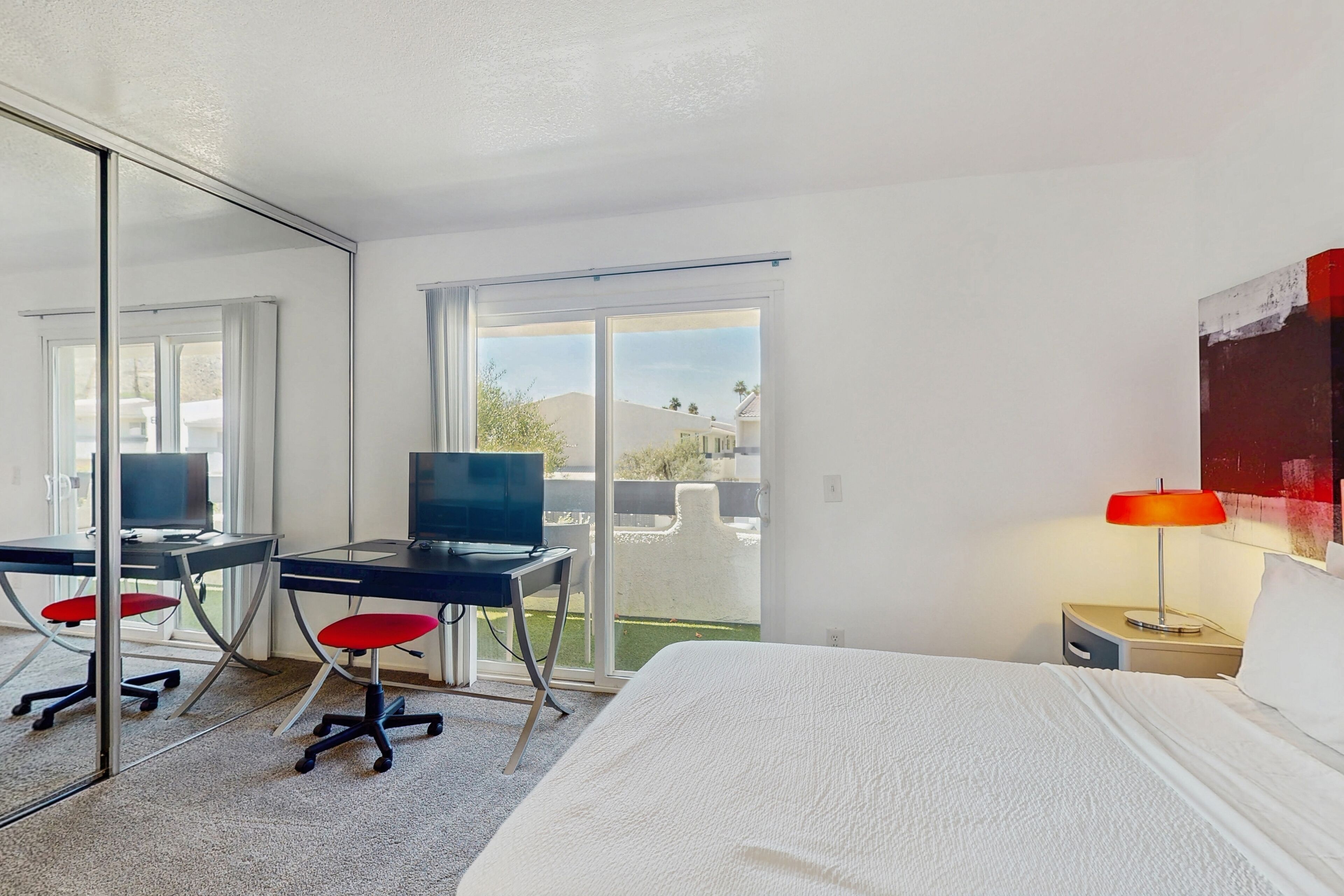 A hotel room with a bed, a desk with a computer, a chair, a lamp, and a sliding glass door leading to a balcony.