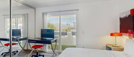 A hotel room with a bed, a desk with a computer, a chair, a lamp, and a sliding glass door leading to a balcony.