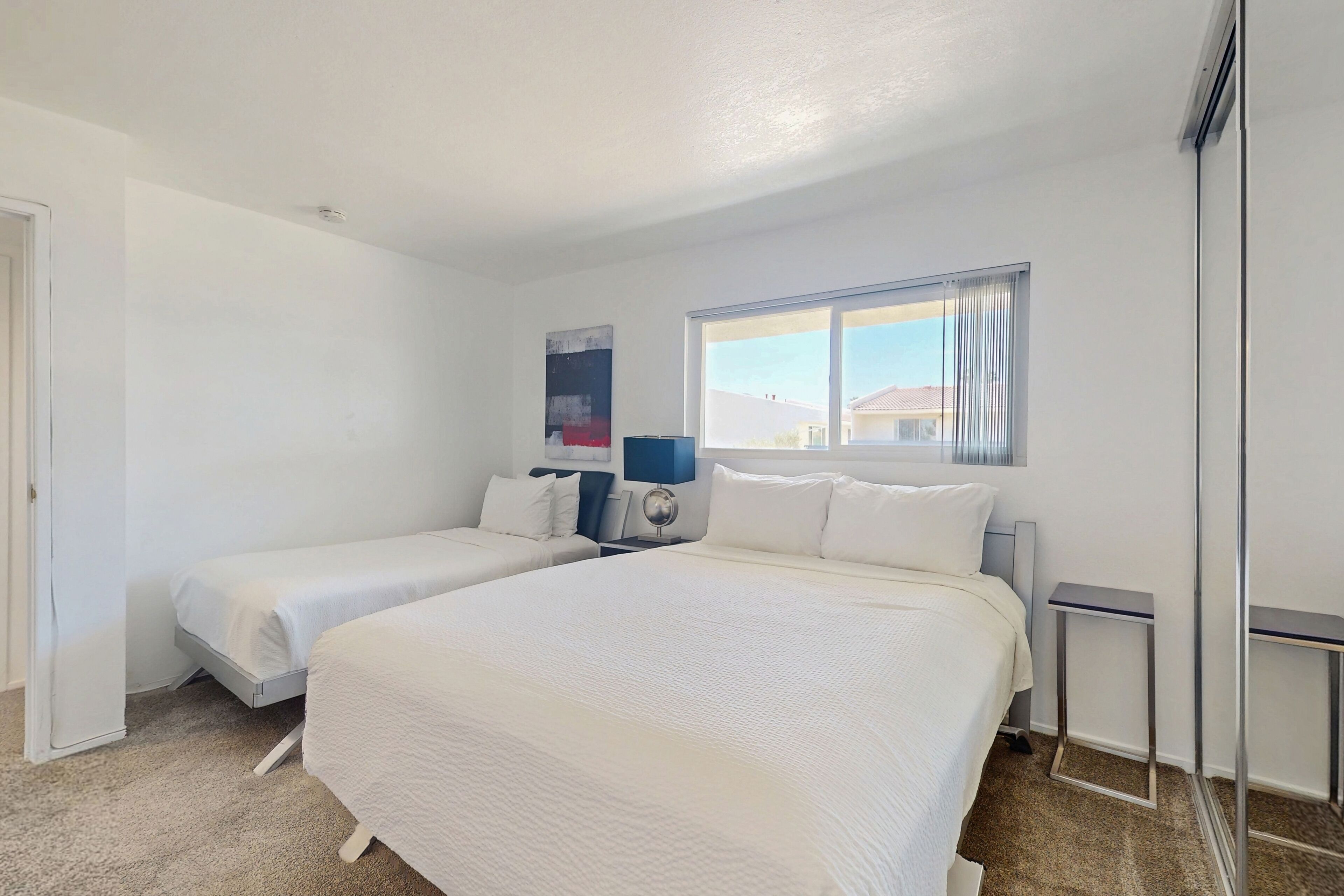 A hotel room with two single beds, a large window with curtains, a bedside table, and a wall-mounted artwork.
