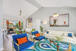 A modern living room with a white sofa, blue armchairs, and a glass coffee table. There is a dining area with a green chair and a white table. A large painting hangs on the wall, and a ceiling fan is visible. - Colorful home with stunning backyard, pool, spa, WiFi, central AC, and fireplace (Palm Springs)