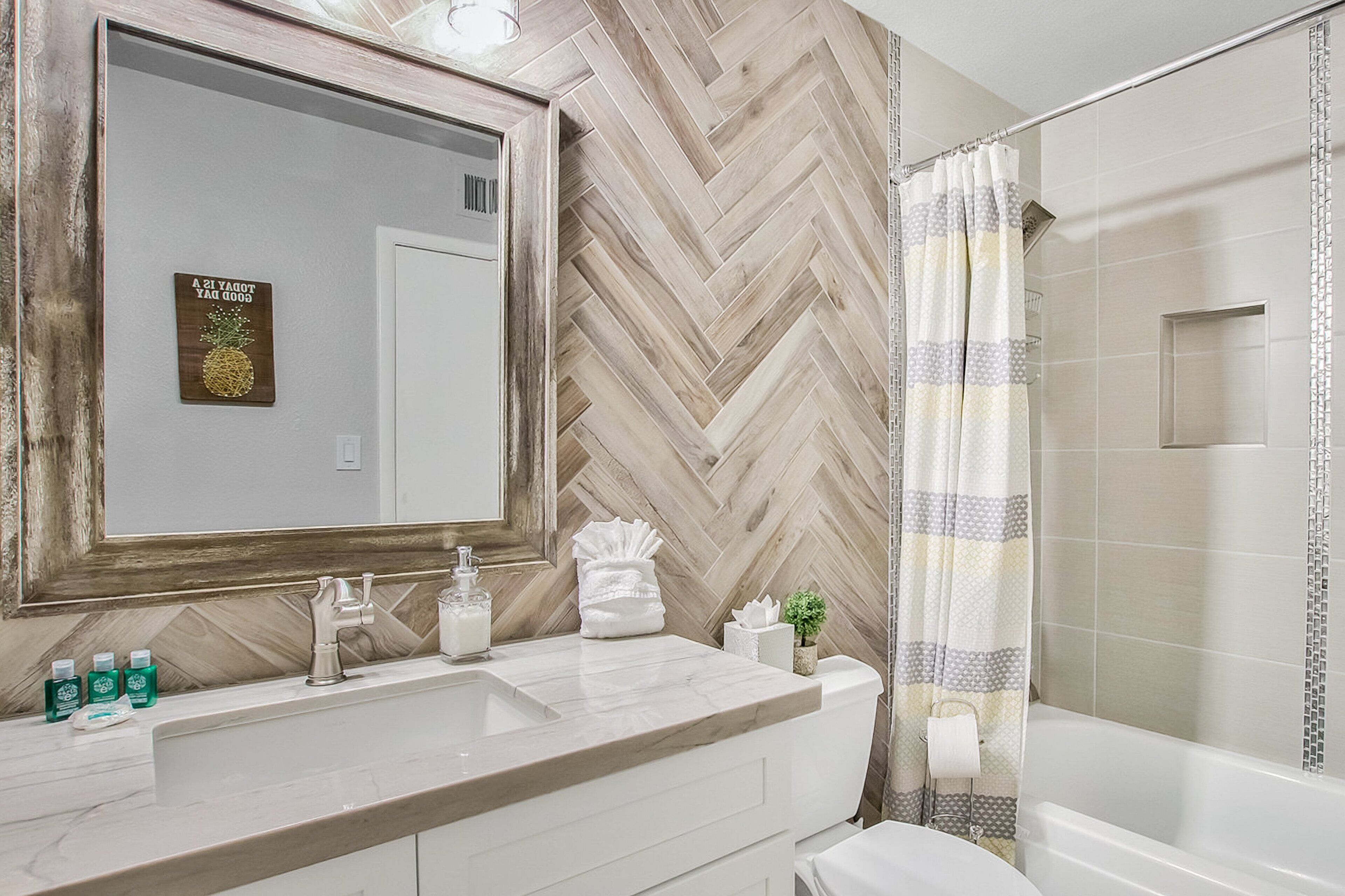 A bathroom with a herringbone wood wall, a large mirror, a white sink with a marble countertop, a glass soap dispenser, and a shower curtain.