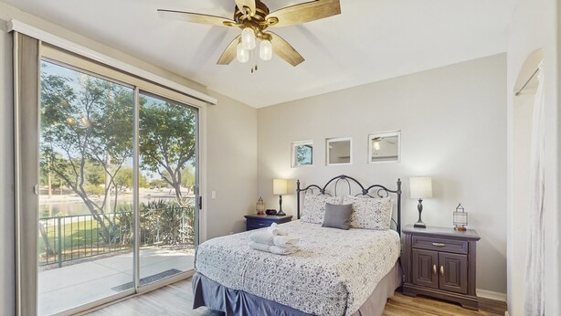 A bedroom with a large sliding glass door leading to a balcony, a bed with a patterned bedspread, two bedside tables with lamps, and a ceiling fan. - Lakefront condo with private washer/dryer, central AC & heated pool (Mesa)