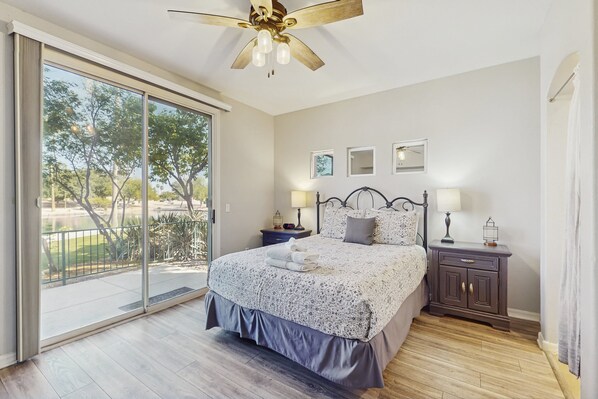 A bedroom with a large sliding glass door leading to a balcony, a bed with a patterned bedspread, two bedside tables with lamps, and a ceiling fan.