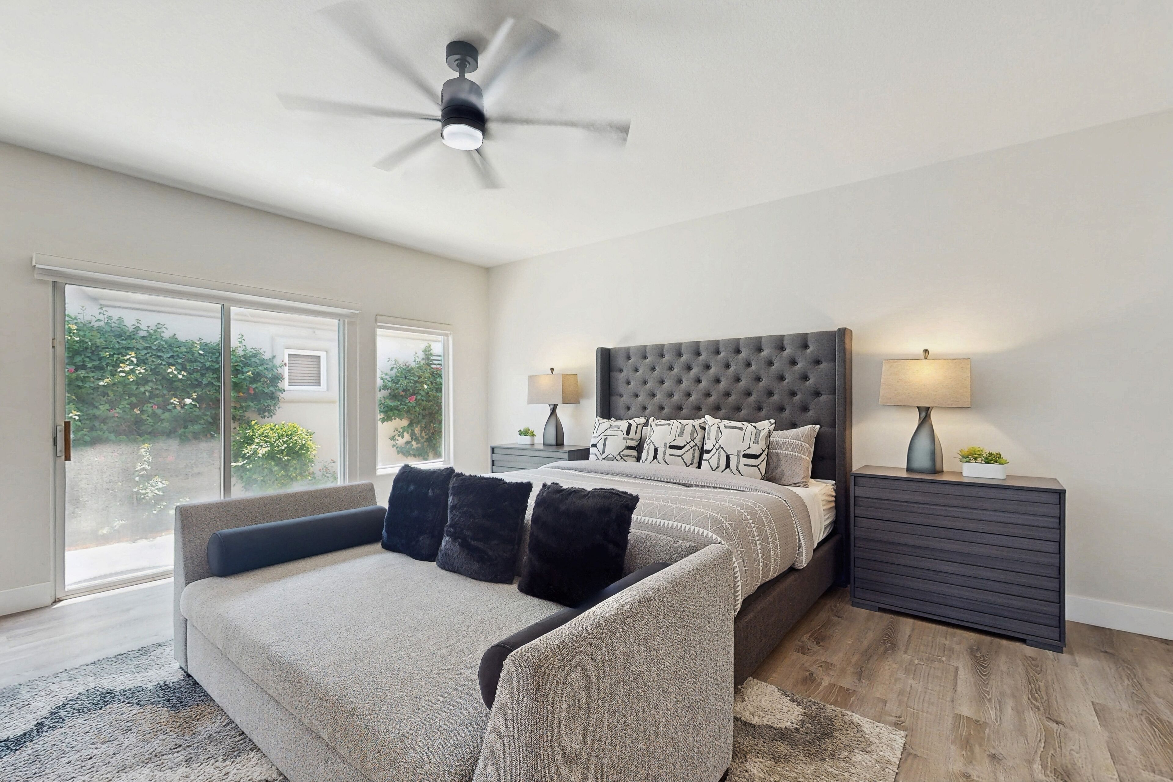 A bedroom with a large bed, a ceiling fan, a nightstand with a lamp, and a view through a sliding door.