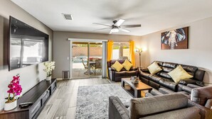 Ein modernes Wohnzimmer mit einem dunkelbraunen Ledersofa, einem grauen Sessel und einem Couchtisch. An der Wand ist ein Flachbildfernseher angebracht, ein Deckenventilator sorgt für Luftzirkulation und ein großes Fenster ist mit Vorhängen versehen.