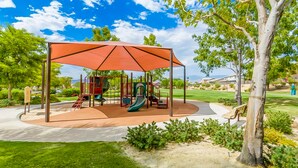 Ein Spielplatz mit überdachtem Spielbereich, Schaukeln und Rutschen, umgeben von Gras und Bäumen.