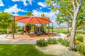 Ein Spielplatz mit überdachtem Spielbereich, Schaukeln und Rutschen, umgeben von Gras und Bäumen.