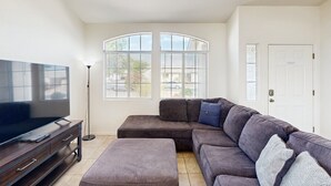 A living room with a sectional sofa, a flat-screen TV, a floor lamp, and a large window with blinds. - Sun-filled dog-friendly home with private pool, putting green, & mountain views (Tucson)