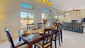 Un comedor con una mesa preparada para cuatro, ventanas amplias, un muro de piedra y una sala de estar con sofá y televisor.