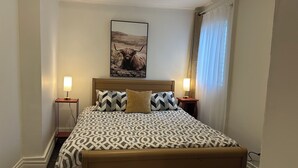 A bedroom with a bed, two bedside tables with lamps, a headboard, a wall-mounted picture, and a window with curtains. - Downtown Digs (St. John's)