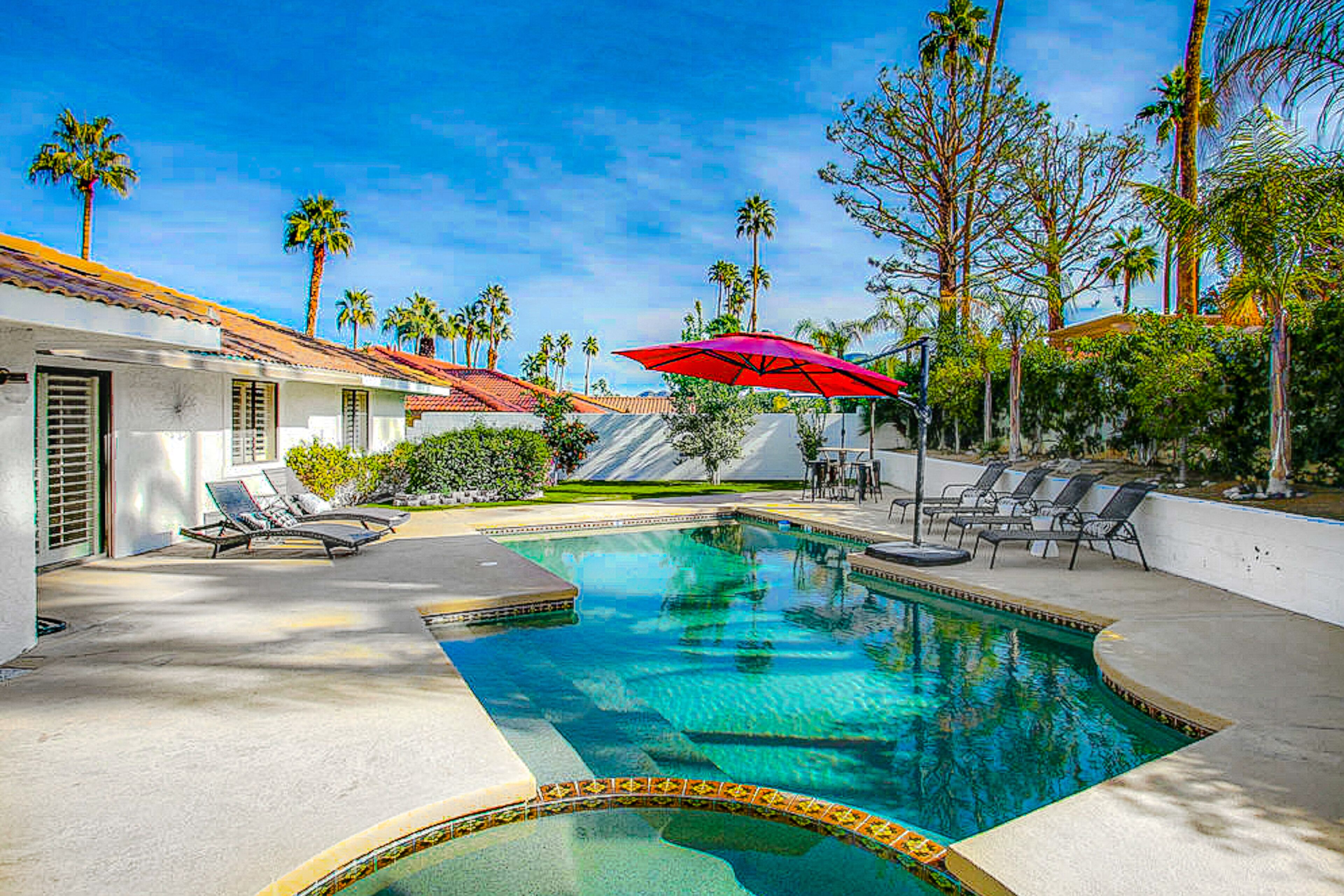 A swimming pool with lounge chairs and umbrellas, surrounded by palm trees and a building with a tiled roof.