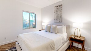 Ein Schlafzimmer mit einem Bett, einem Fenster, einem Wandbild und einem Nachttisch mit einer Lampe.