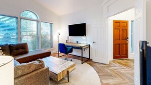 Ein Wohnzimmer mit Sofa, Couchtisch und Fernseher. Es gibt einen Schreibtisch mit Stuhl und Computer. Das Zimmer hat große Fenster und eine Tür.