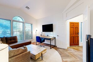 Ein Wohnzimmer mit Sofa, Couchtisch und Fernseher. Es gibt einen Schreibtisch mit Stuhl und Computer. Das Zimmer hat große Fenster und eine Tür.