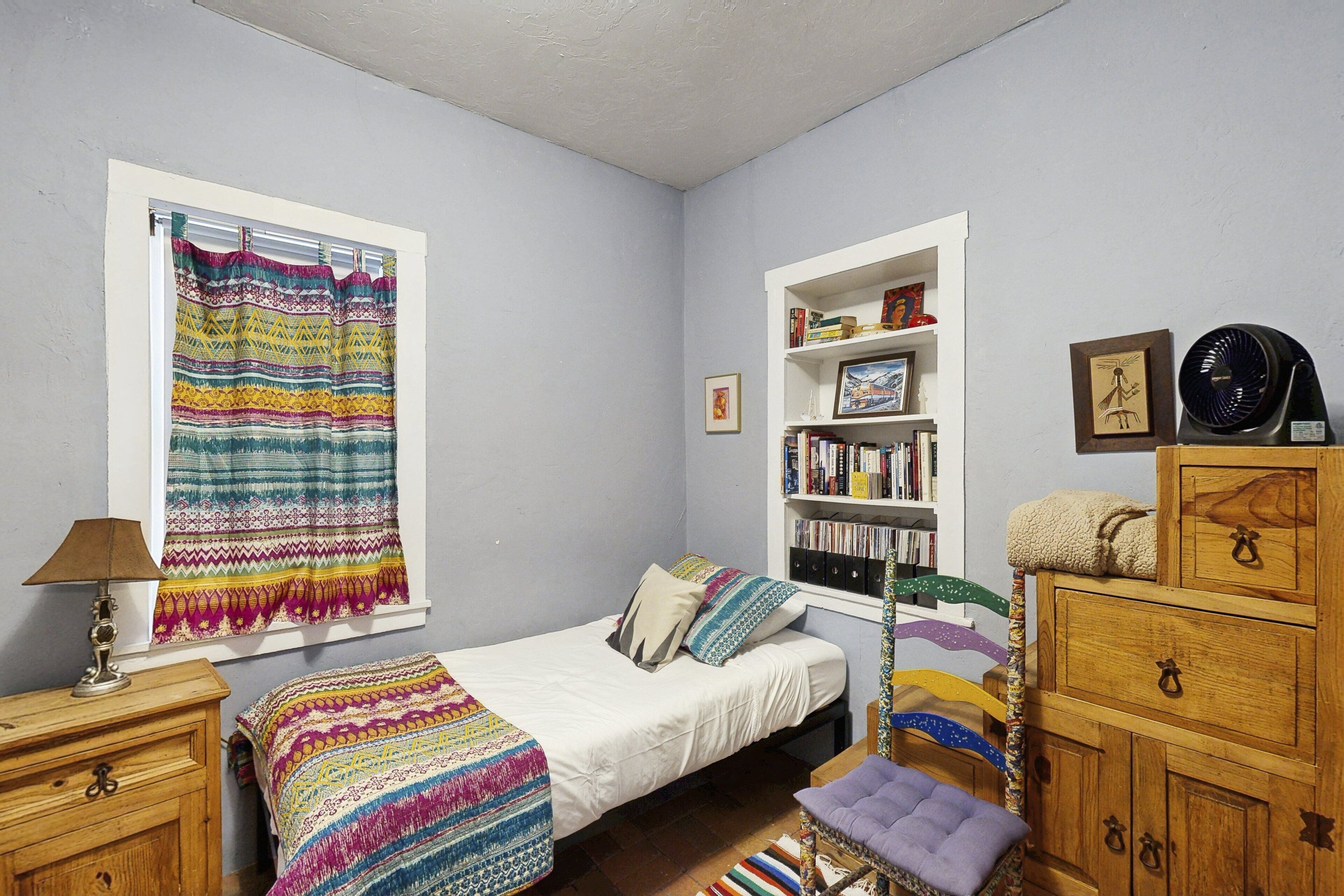 Un dormitorio pequeño con cama, ventana con cortinas, estantería, cómoda de madera y escritorio con lámpara.