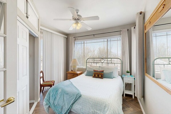 Un dormitorio con cama, mesita de noche, silla, ventana con persianas y ventilador de techo.