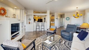 A living room with a TV, seating, a fireplace, and a kitchen in the background. - Beautiful first-floor condo with pool, hot tub, & full kitchen - near hiking (Oro Valley)