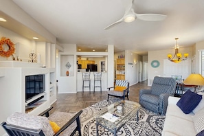 A living room with a TV, seating, a fireplace, and a kitchen in the background. - Beautiful first-floor condo with pool, hot tub, & full kitchen - near hiking (Oro Valley)