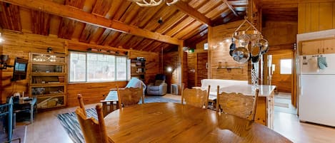 Interior de una cabaña de madera con una gran mesa de comedor, sillas, una araña y una zona de cocina.