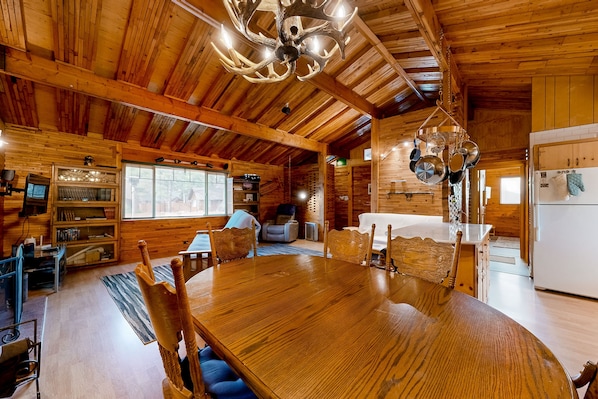 L’intérieur d’un chalet en bois comprend une grande table à manger, des chaises, un lustre et un coin cuisine.