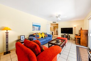 Ein Wohnzimmer mit blauem Sofa, roten Sesseln, einem Couchtisch, einem Fernseher und einem Deckenventilator.
