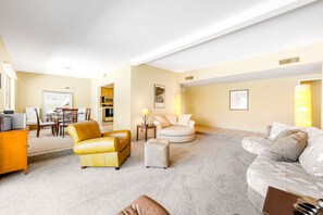 Ein geräumiges Wohnzimmer mit einem gelben Sofa, einem beigen Sessel und einem gemusterten Sofa. Ein Essbereich mit Tisch und Stühlen ist vorhanden, und im Hintergrund befindet sich eine Küche.
