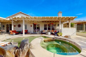 A pool area with a covered patio, seating, and a large rock feature. - Luxe and spacious retreat with a pool, pool spa, firepit, grill, game room, & AC (Indio)