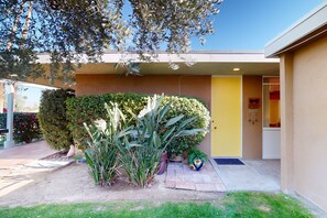 Un bâtiment moderne avec une porte jaune, un jardin aménagé avec différentes plantes et un ciel dégagé.