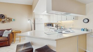 Una cocina moderna con una isla central, muebles blancos y un microondas. Hay un sofá de cuero marrón y una mesita de madera con una lámpara.