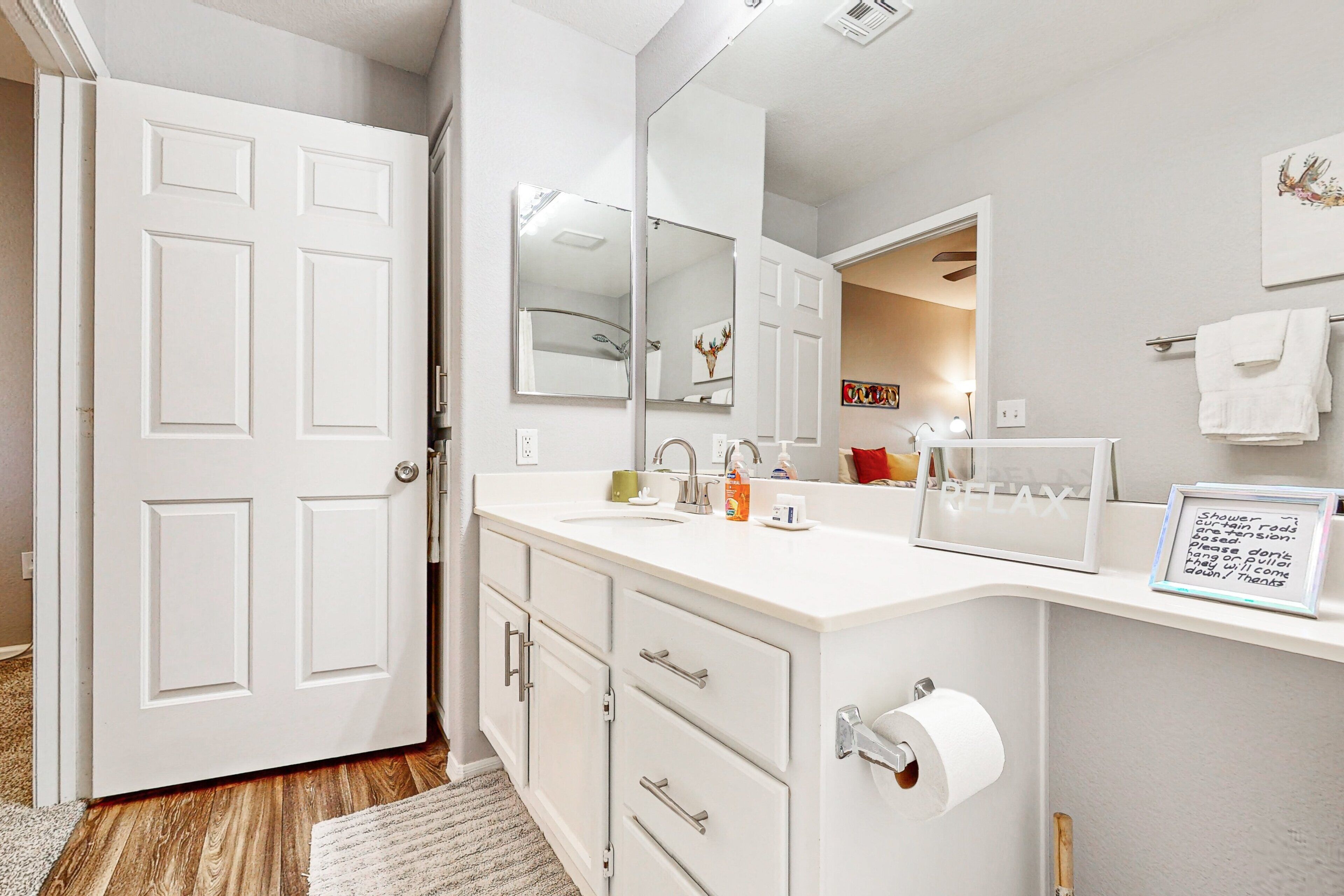 A bathroom with a white vanity, a large mirror, a towel rack, and a framed note on the wall.