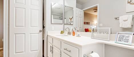 A bathroom with a white vanity, a large mirror, a towel rack, and a framed note on the wall.
